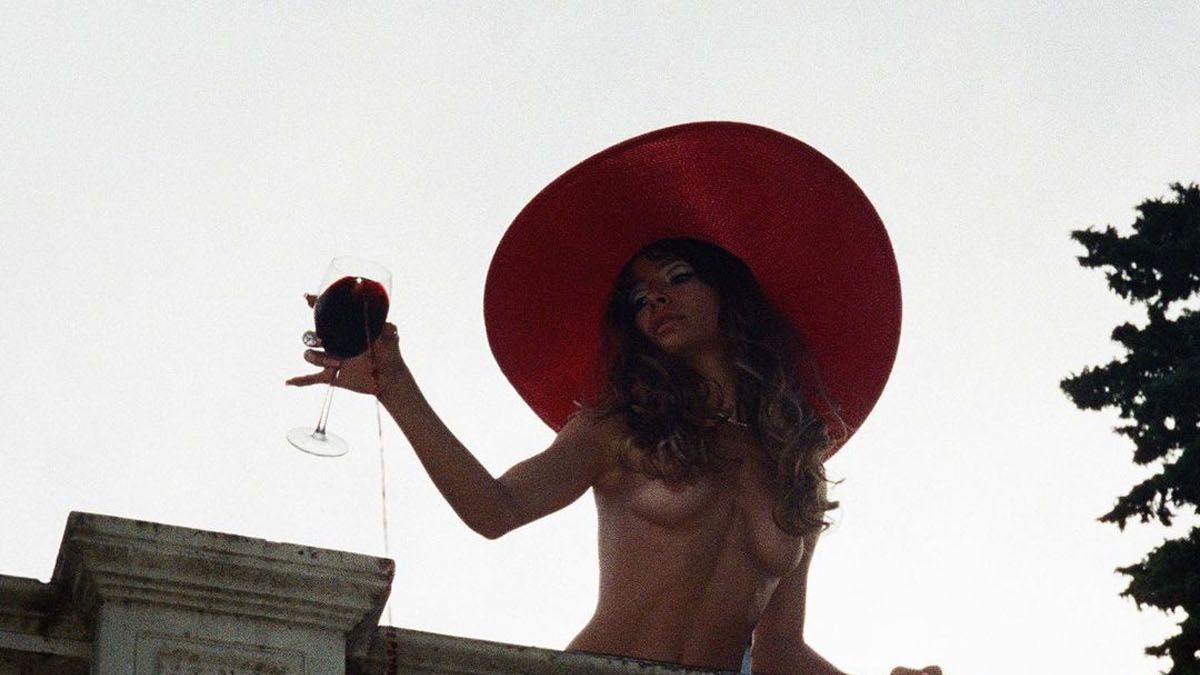 Brenda Asnicar, con sombrero, una copa de vino... y nada más