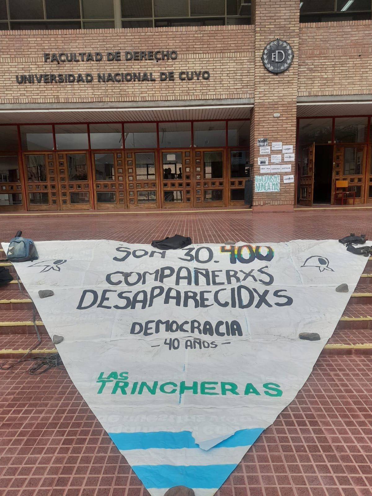 Bandera en forma de pañuelo de Madres de Plaza de Mayo colocada en el ingreso a la Faclutad de Derecho de la UNCuyo. Bandera en forma de pañuelo de Madres de Plaza de Mayo colocada en el ingreso a la Faclutad de Derecho de la UNCuyo.