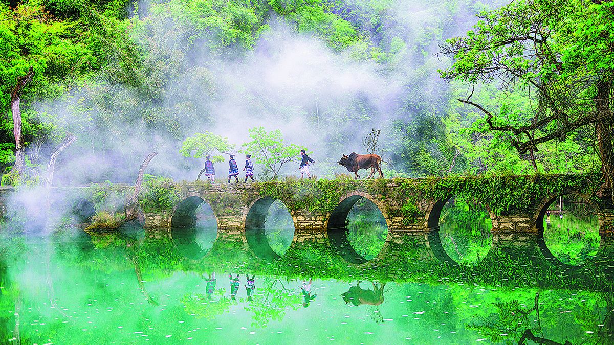 Unos aldeanos cruzan un puente de piedra envuelto por la niebla de la mañana en el condado de Libo